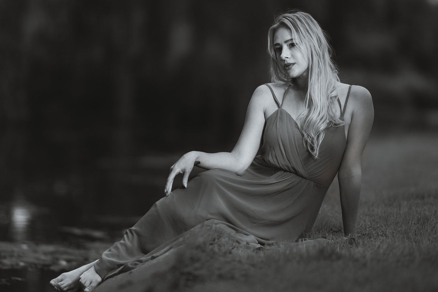 Black and white photo of a blonde woman in a flowing dress sitting gracefully on grass outdoors.