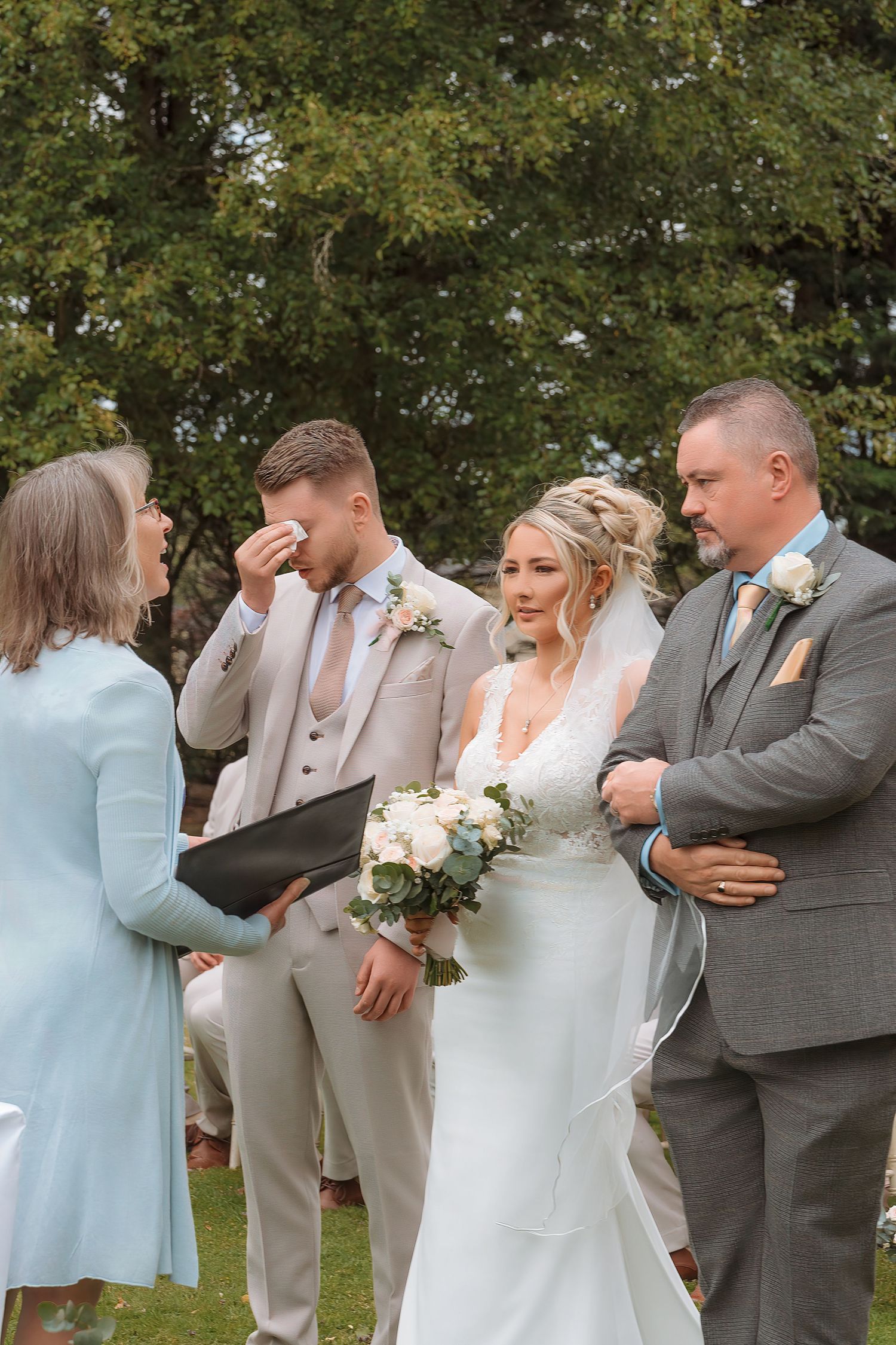 Emotional groom wiping tears during outdoor wedding ceremony with bride holding white bouquet and wedding party.