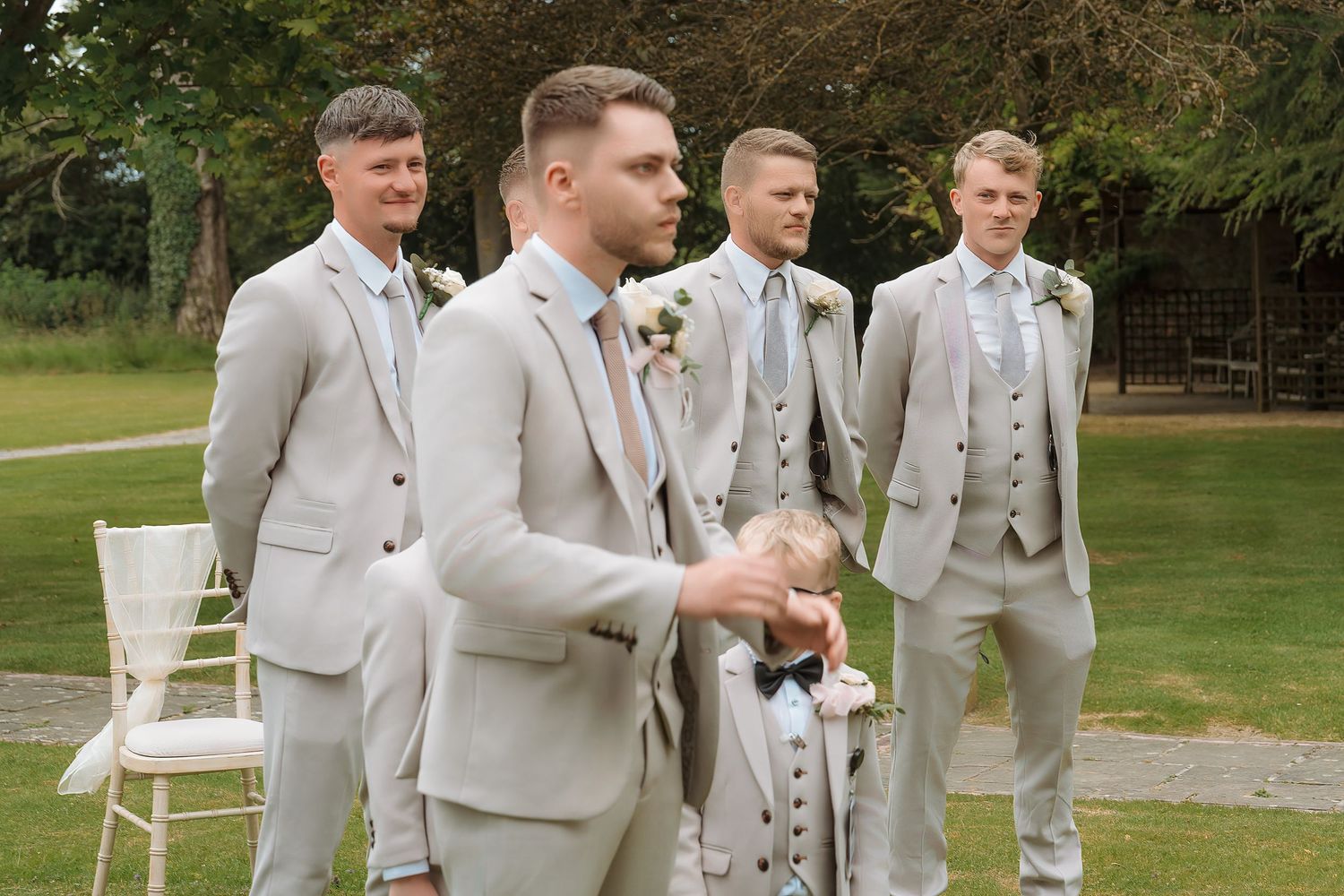 Groomsmen in matching light grey suits standing outdoors at a wedding ceremony on a green lawn.