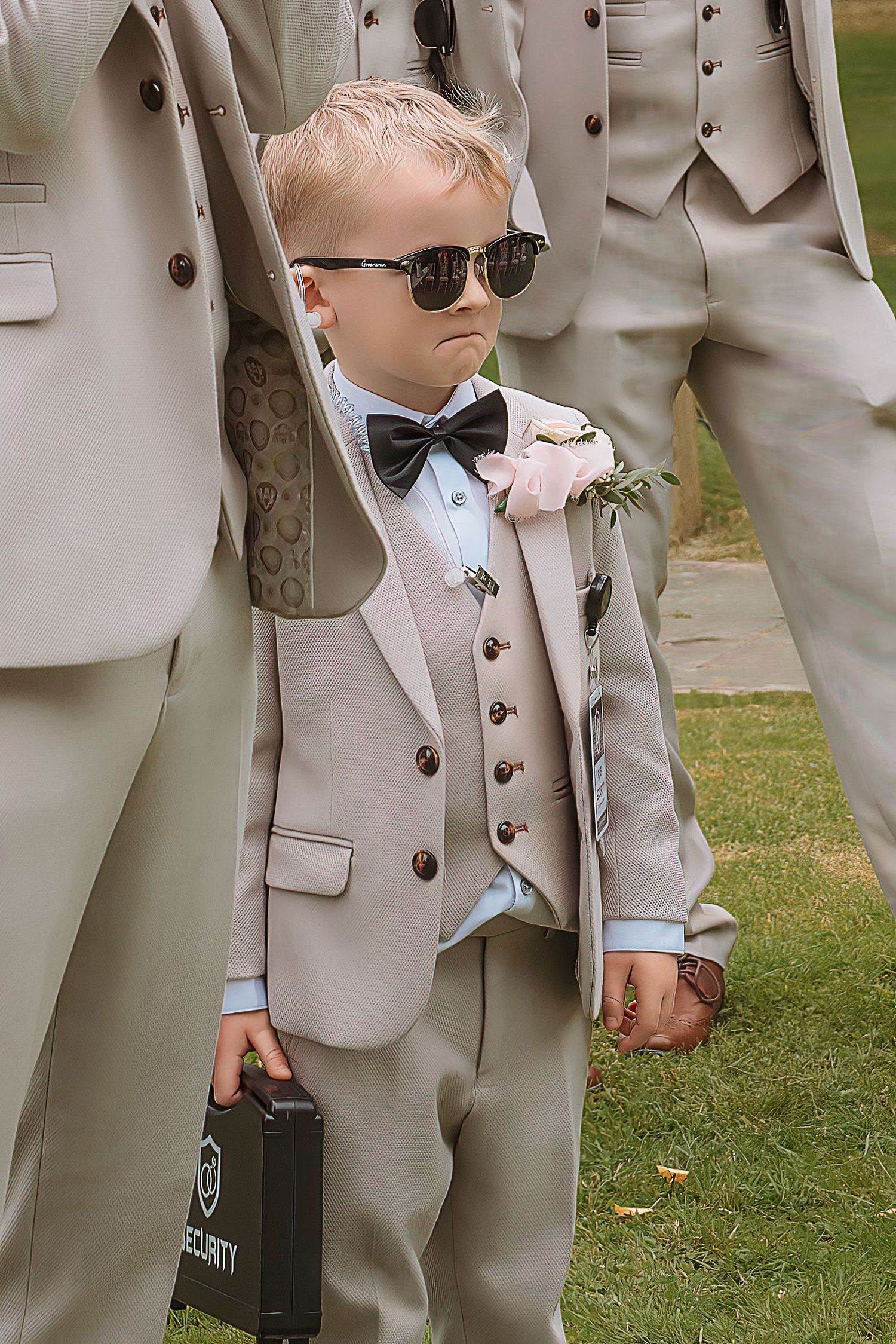 Adorable boy in tan suit, bow tie, sunglasses, holding security case at wedding.