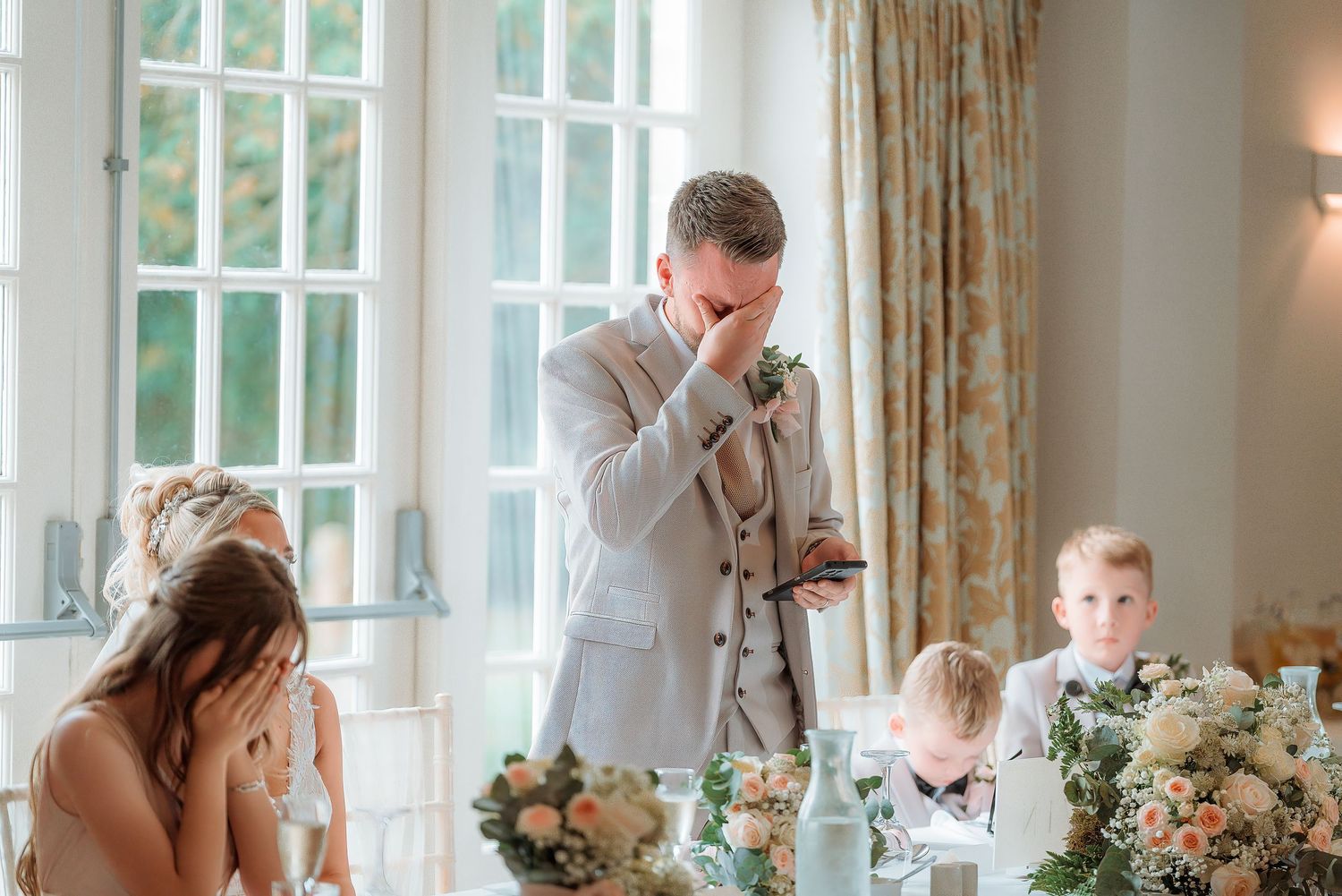 Emotional groom wiping tears during wedding speech at elegant venue with floral centerpieces and young guests seated nearby.