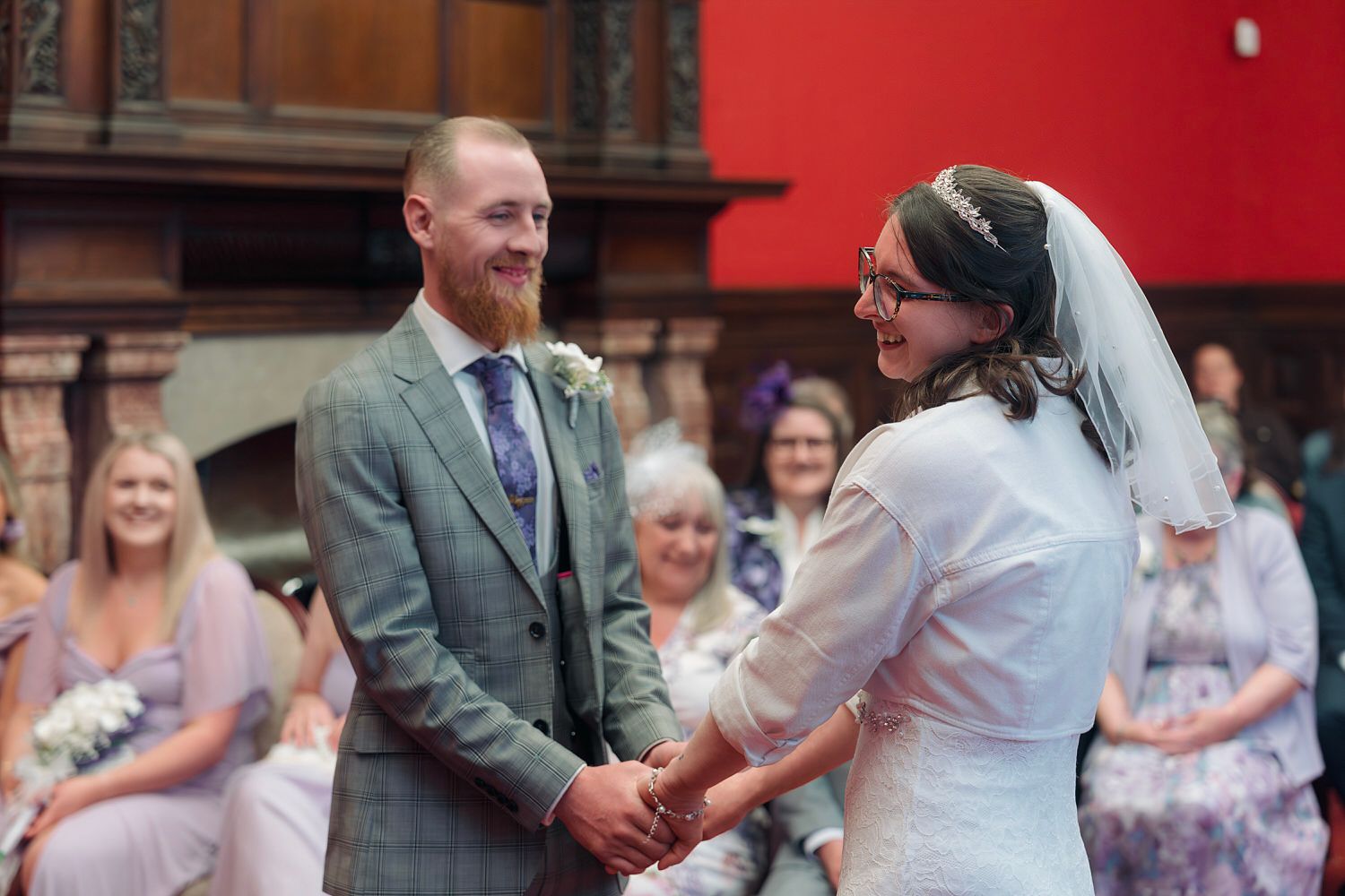 Bride and groom holding hands and smiling during wedding ceremony in elegant venue with guests seated behind.