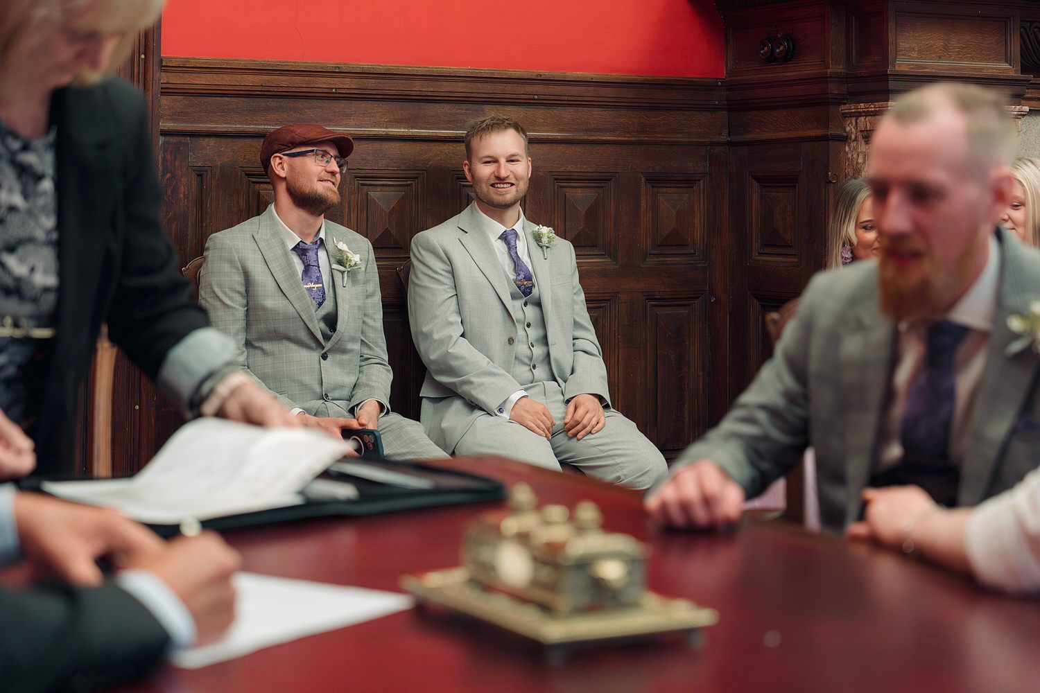 Two grooms in grey suits smile during their wedding ceremony at a wooden-paneled registry office.