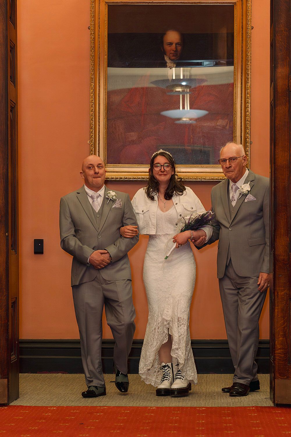 Bride in white lace dress walks with two men in gray suits under a portrait painting at a wedding ceremony.