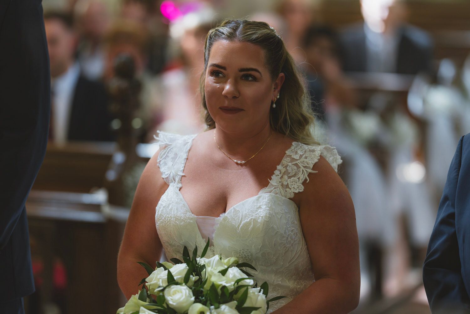 Bride in lace wedding dress holding white bouquet walks down church aisle, smiling radiantly.