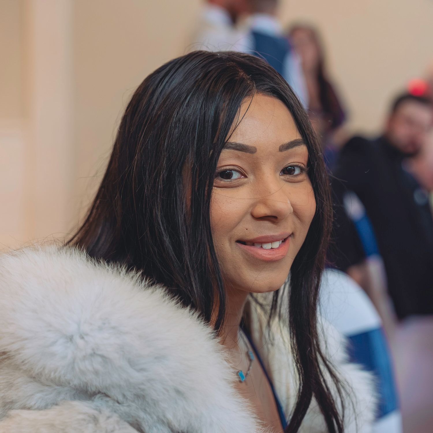A young woman with long black hair smiles warmly, wearing a white fur coat with a turquoise necklace at an indoor event.