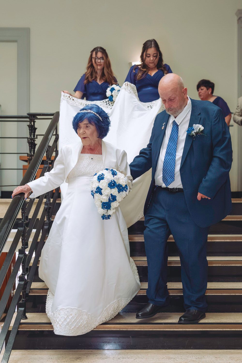 Elderly bride in white gown and blue hat descends stairs with groom in blue suit, bridesmaids holding train behind them.
