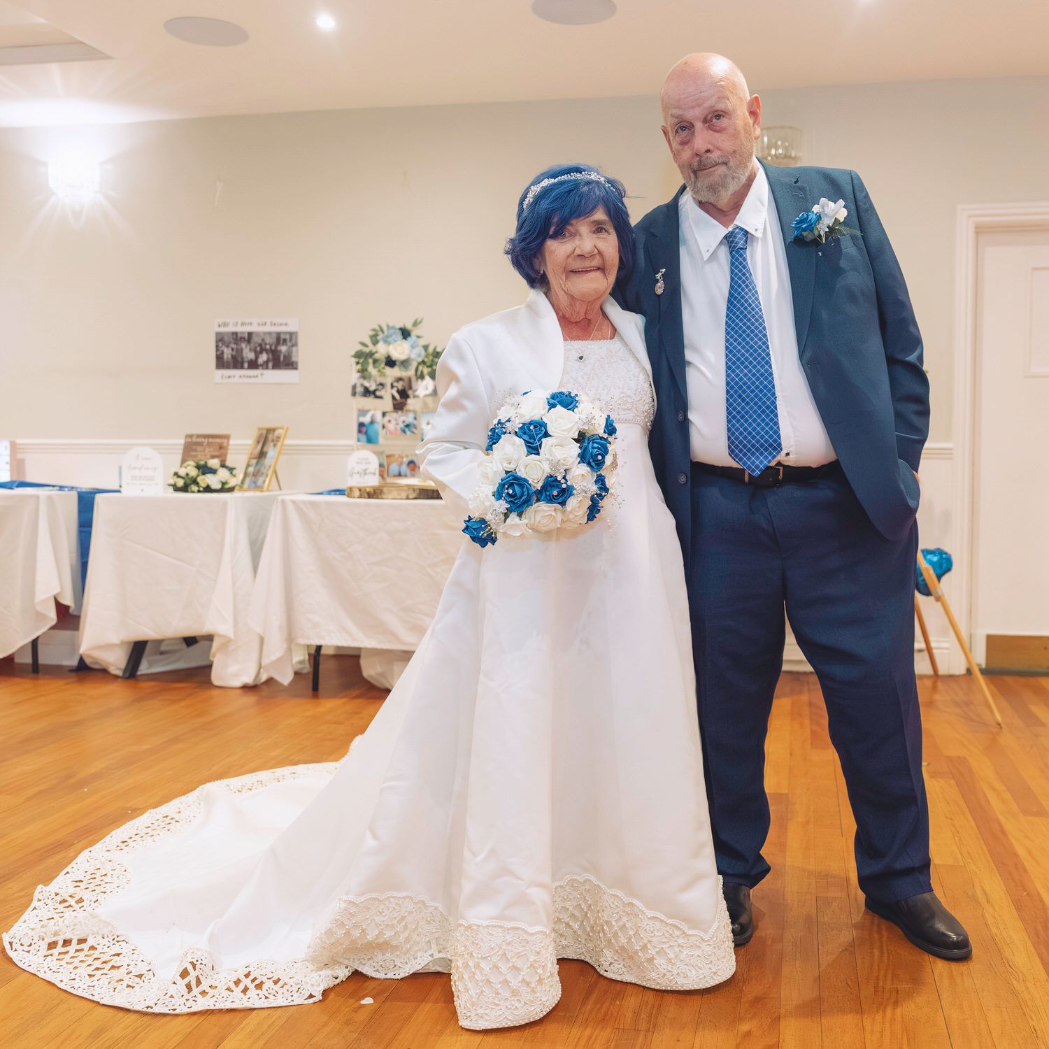 Elderly couple in wedding attire, bride in white gown with blue bouquet, groom in navy suit, posing in decorated reception hall.