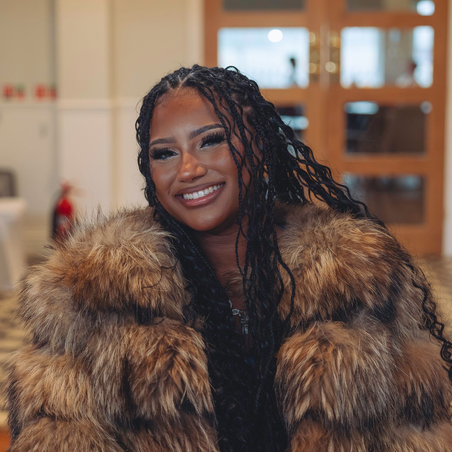Smiling woman with braids wearing a luxurious fur coat indoors, exuding confidence and style.