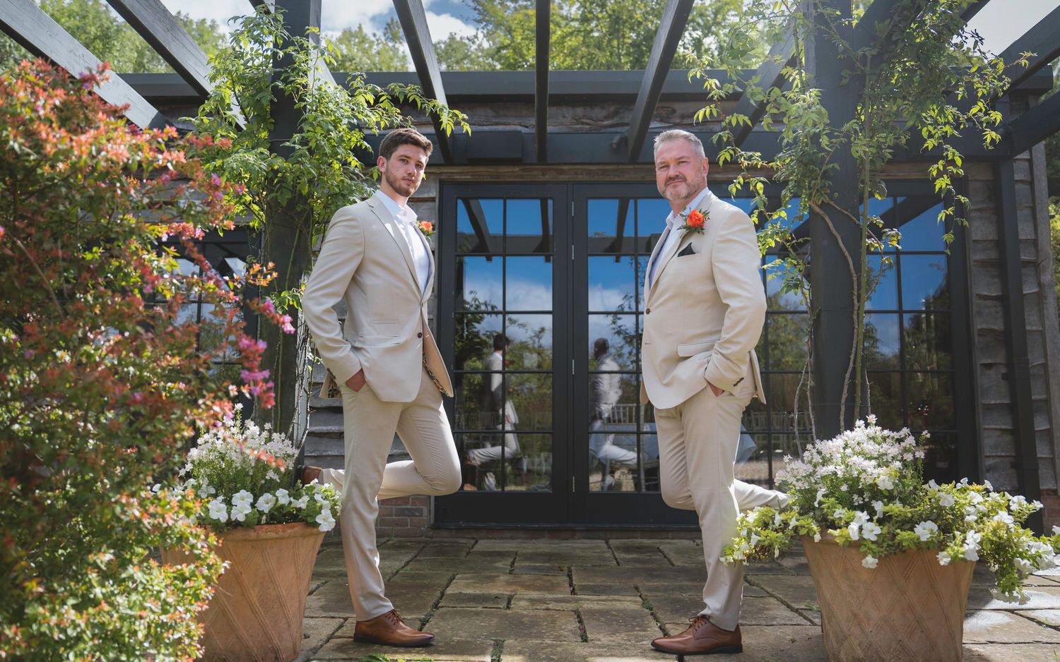 Two grooms in matching beige suits pose outdoors at a garden wedding venue surrounded by lush greenery and flowers.