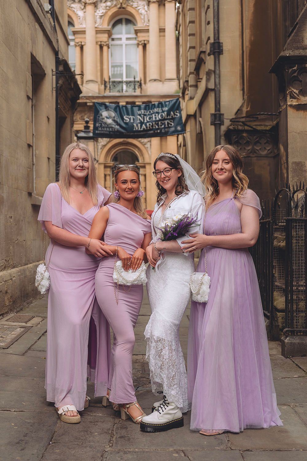 Bride in white lace dress with three bridesmaids in lavender gowns posing at St. Nicholas Markets, Bristol.
