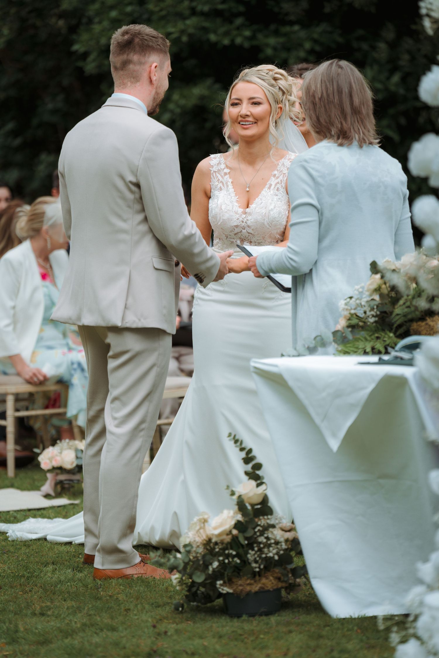 Bride and groom holding hands during outdoor wedding ceremony, bride smiling in white mermaid dress, groom in beige suit.