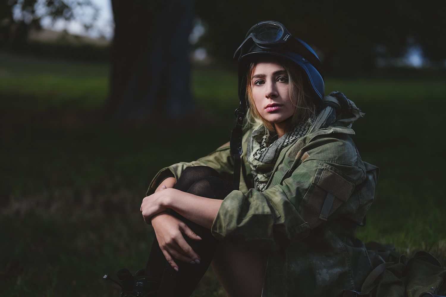 Young person in camouflage military outfit and helmet sitting on grass in a park, looking thoughtful.