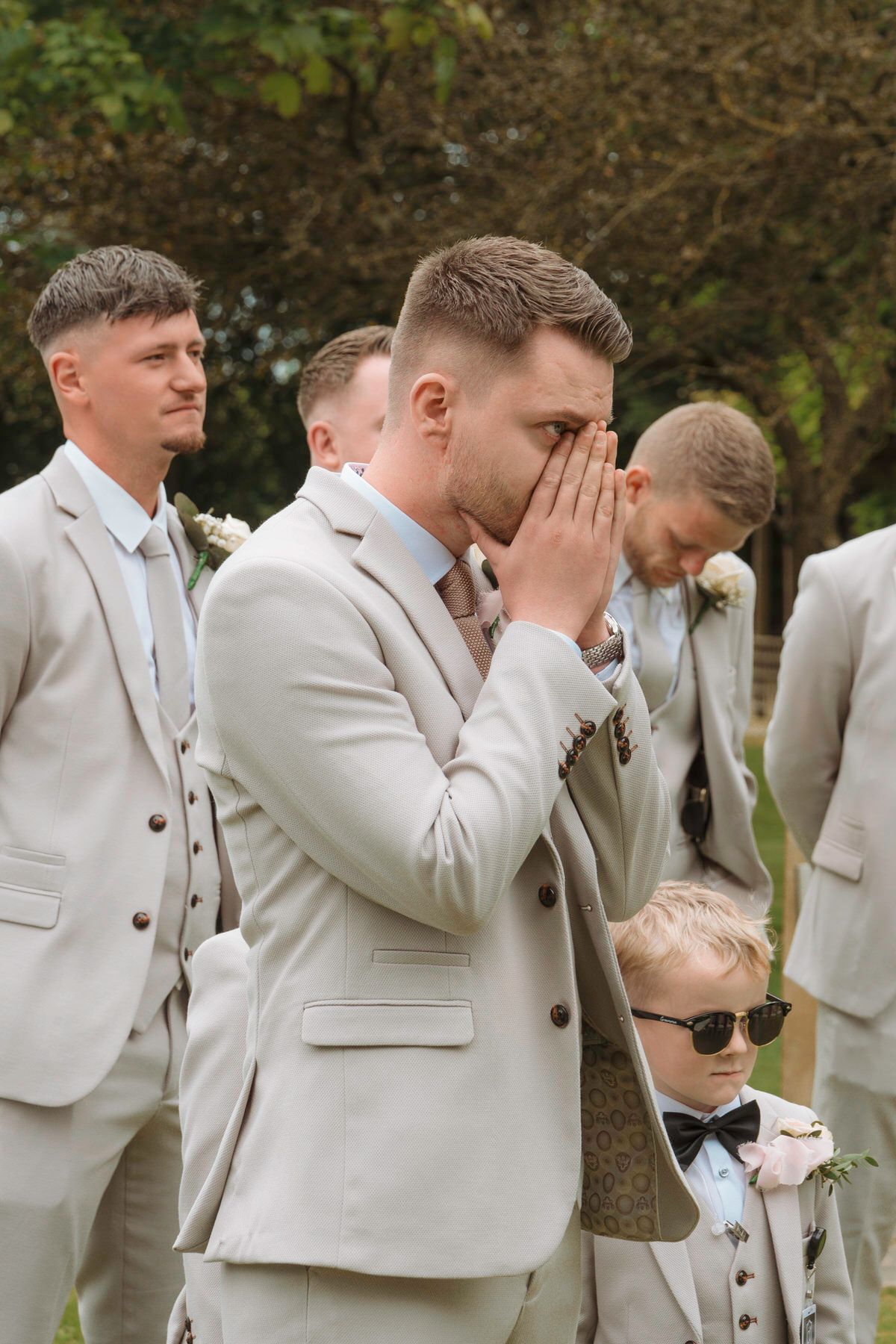 Emotional groom covering face with hands while groomsmen stand behind him outdoors.