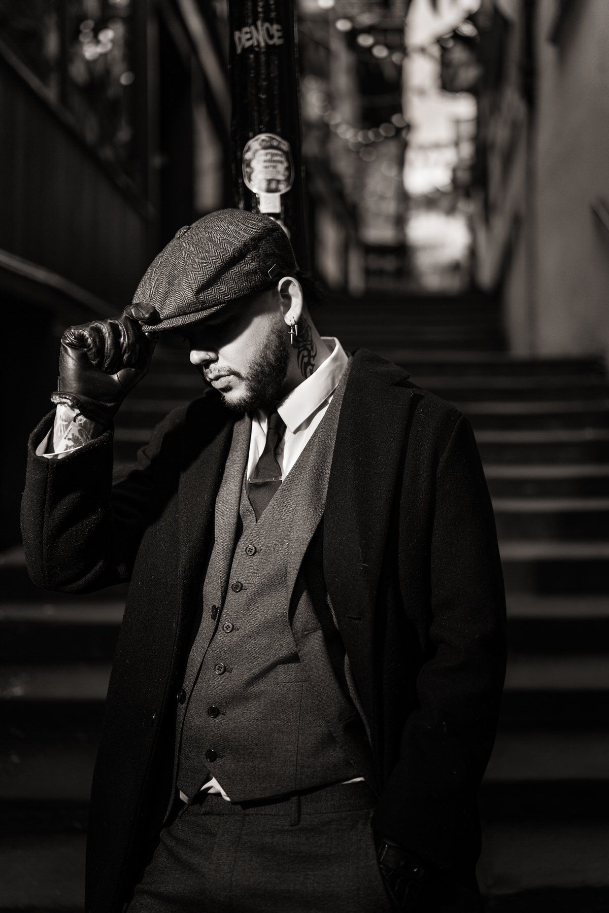 Black and white portrait of stylish man in suit vest adjusting flat cap on urban stairs.