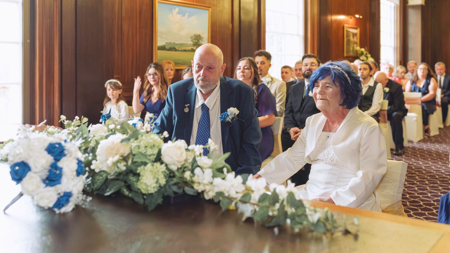 Elderly couple signing wedding documents at a formal ceremony, surrounded by guests and white floral arrangements.
