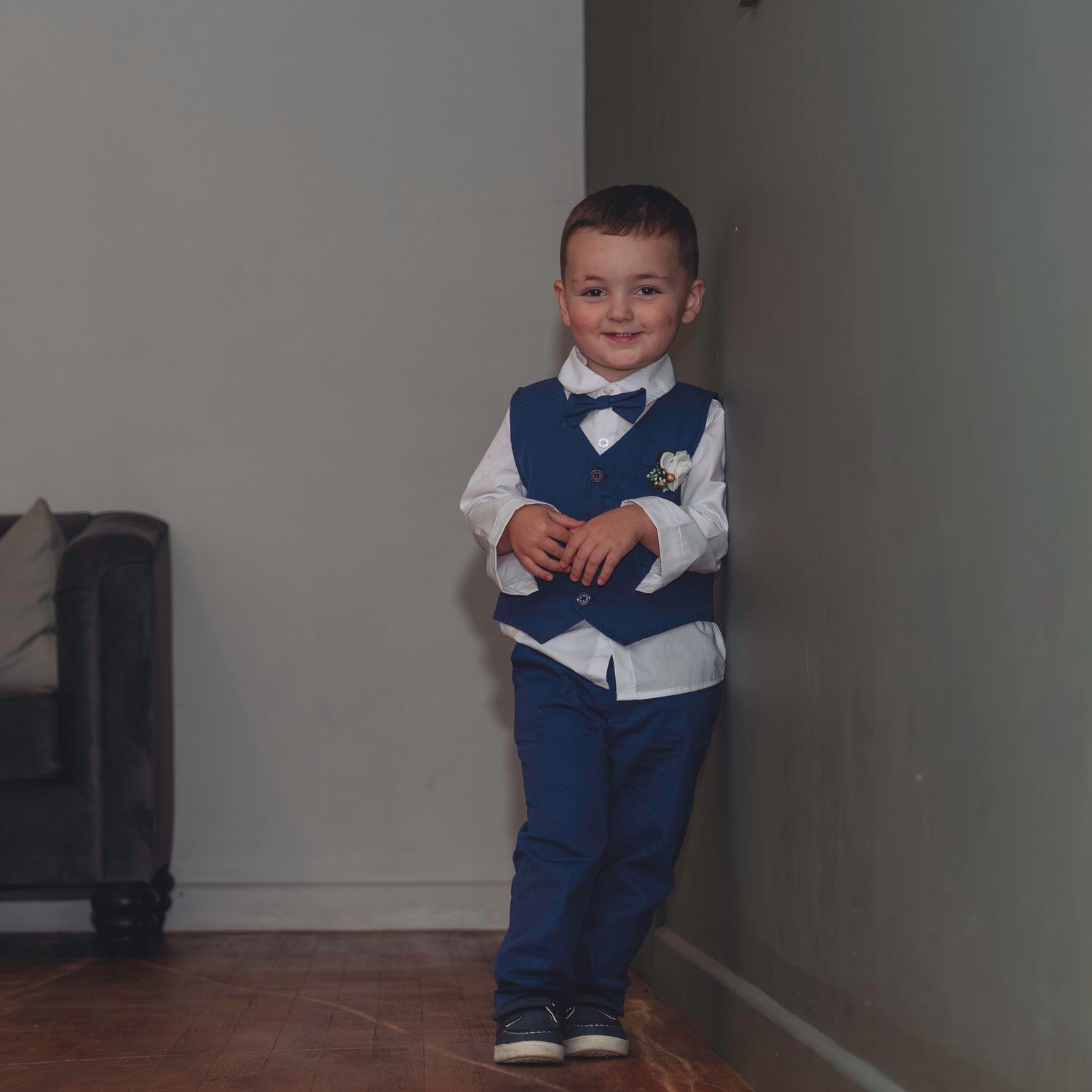 Young boy in blue vest, bow tie, and trousers standing proudly in a corner, dressed in formal attire.