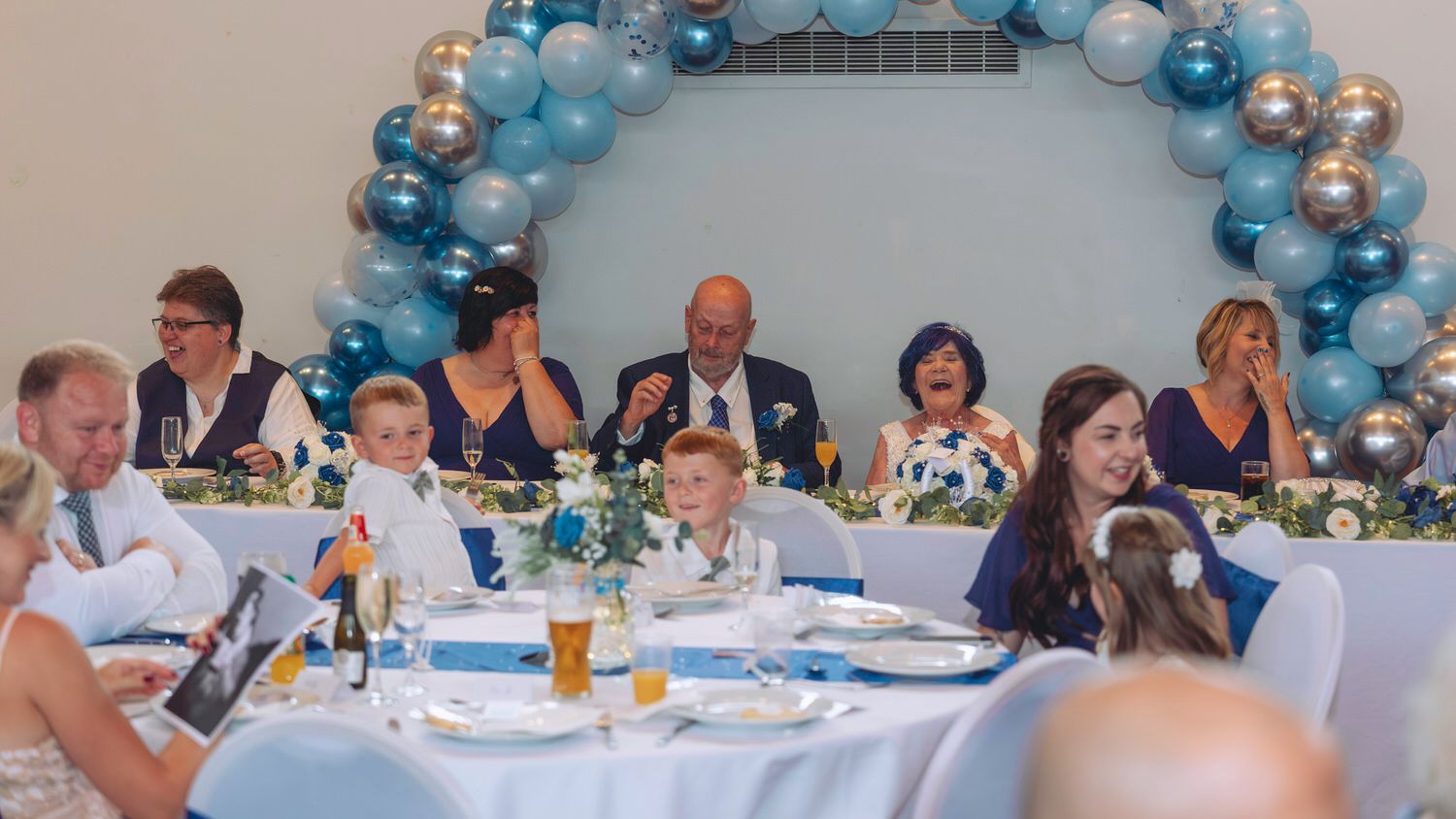 Wedding reception guests seated at decorated tables with blue and white balloon arch backdrop.