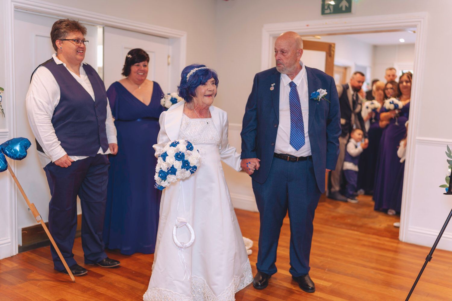 Elderly bride in white gown holds bouquet walking with groom in blue suit at wedding ceremony with guests watching.