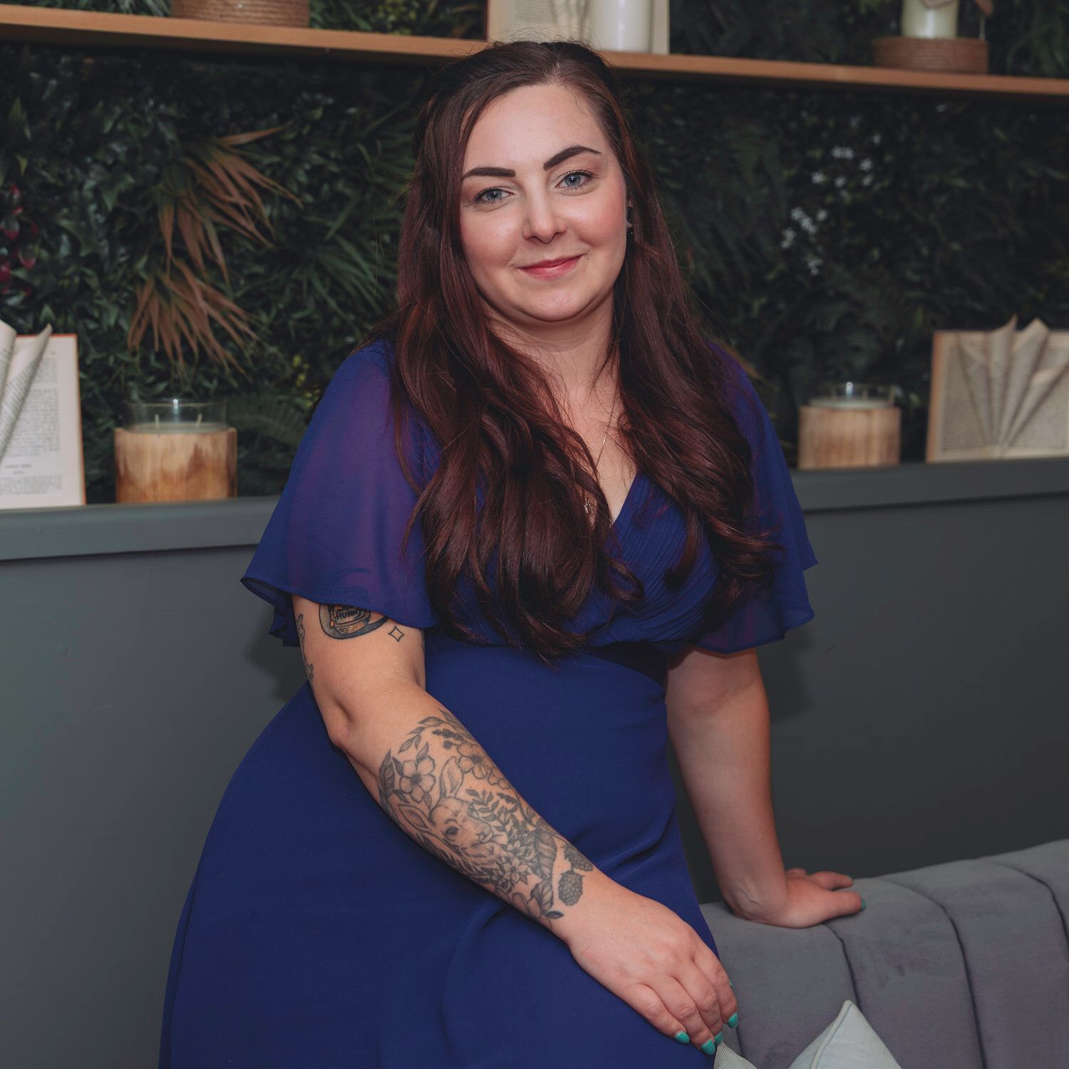 Woman in royal blue wrap dress with floral arm tattoo seated in elegant venue with greenery backdrop.