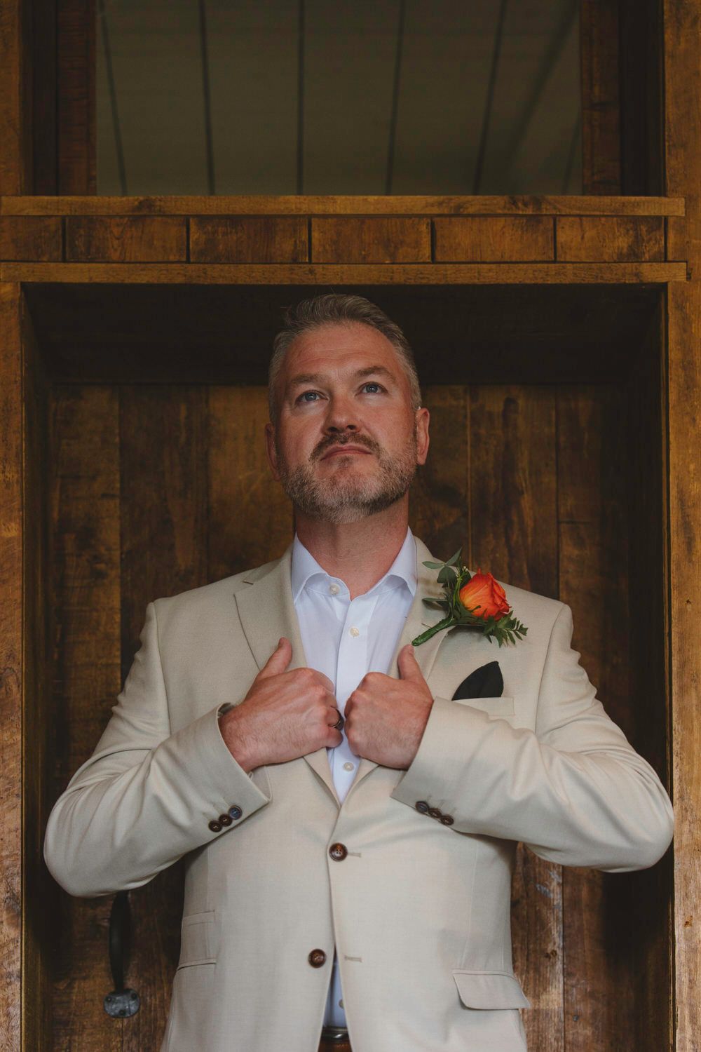 Groom in cream blazer with orange boutonniere adjusts jacket lapels, standing in rustic wooden doorframe.