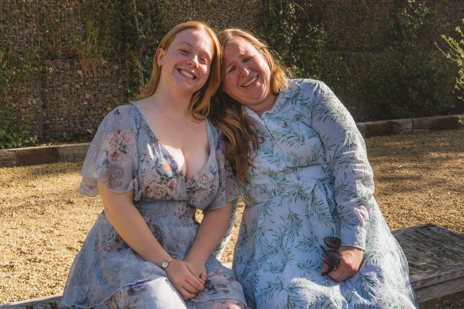 Two women in floral blue dresses smile together outdoors on a sunny day.