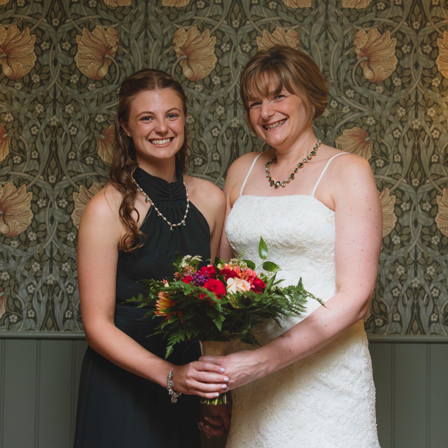 Two women pose together at a wedding, one in a white bridal gown holding a bouquet, the other in a black dress.
