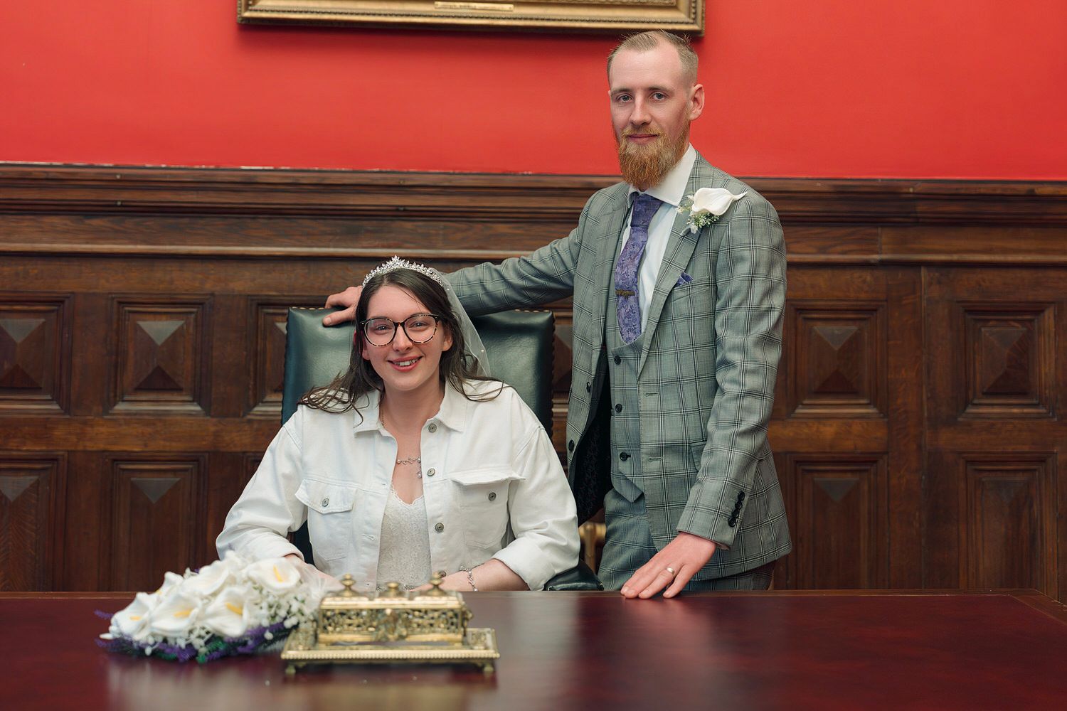 Couple posing at wedding registry signing table in elegant red-walled room, bride seated in white jacket, groom standing beside her.
