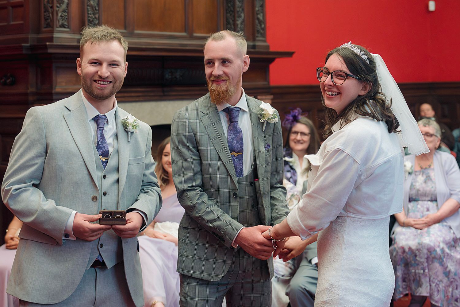 Wedding ceremony with two grooms in grey suits and bride in white dress exchanging rings, smiling guests in background.
