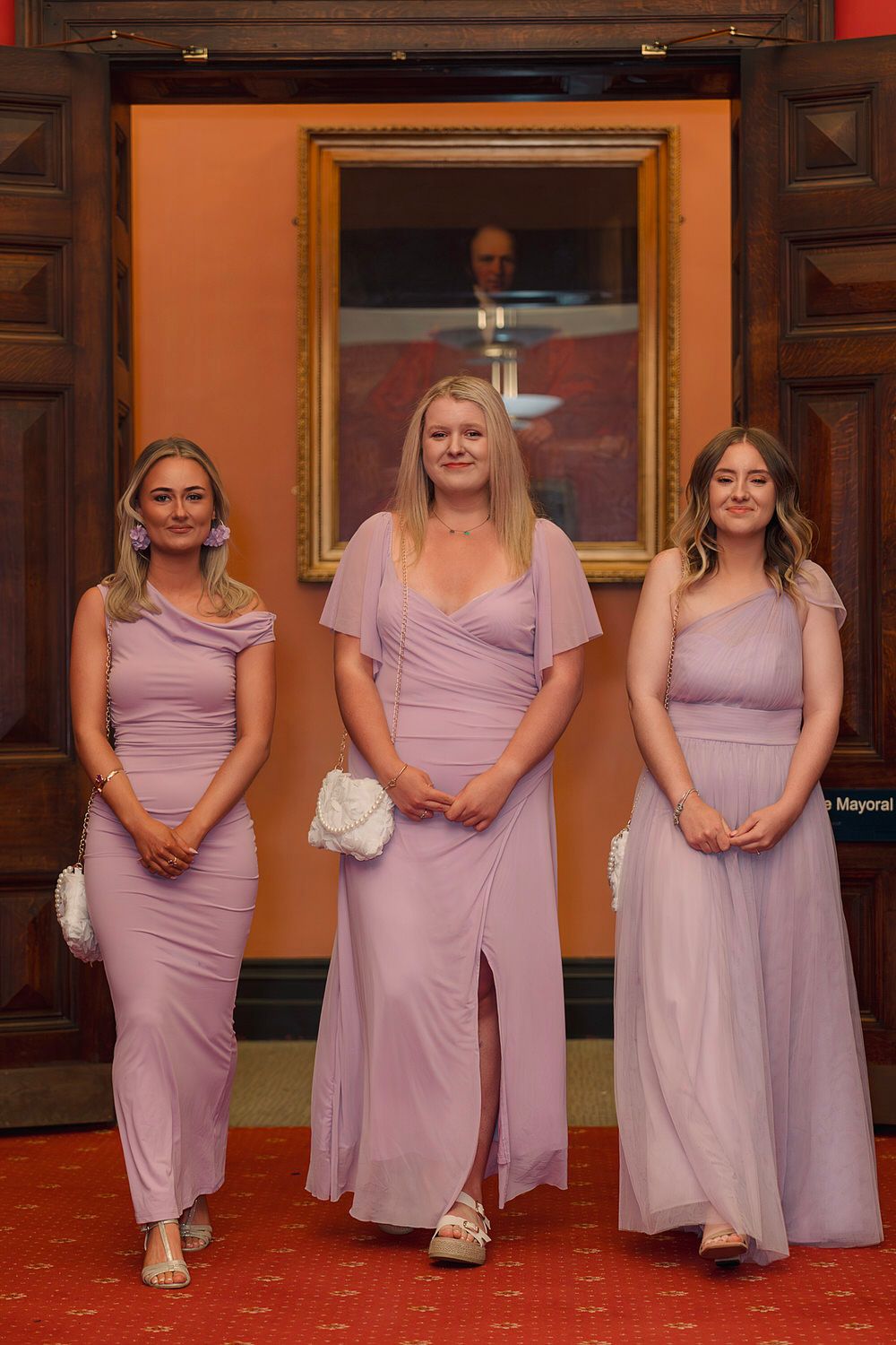 Three bridesmaids in matching lavender gowns pose elegantly in a grand hall with ornate wooden decor.
