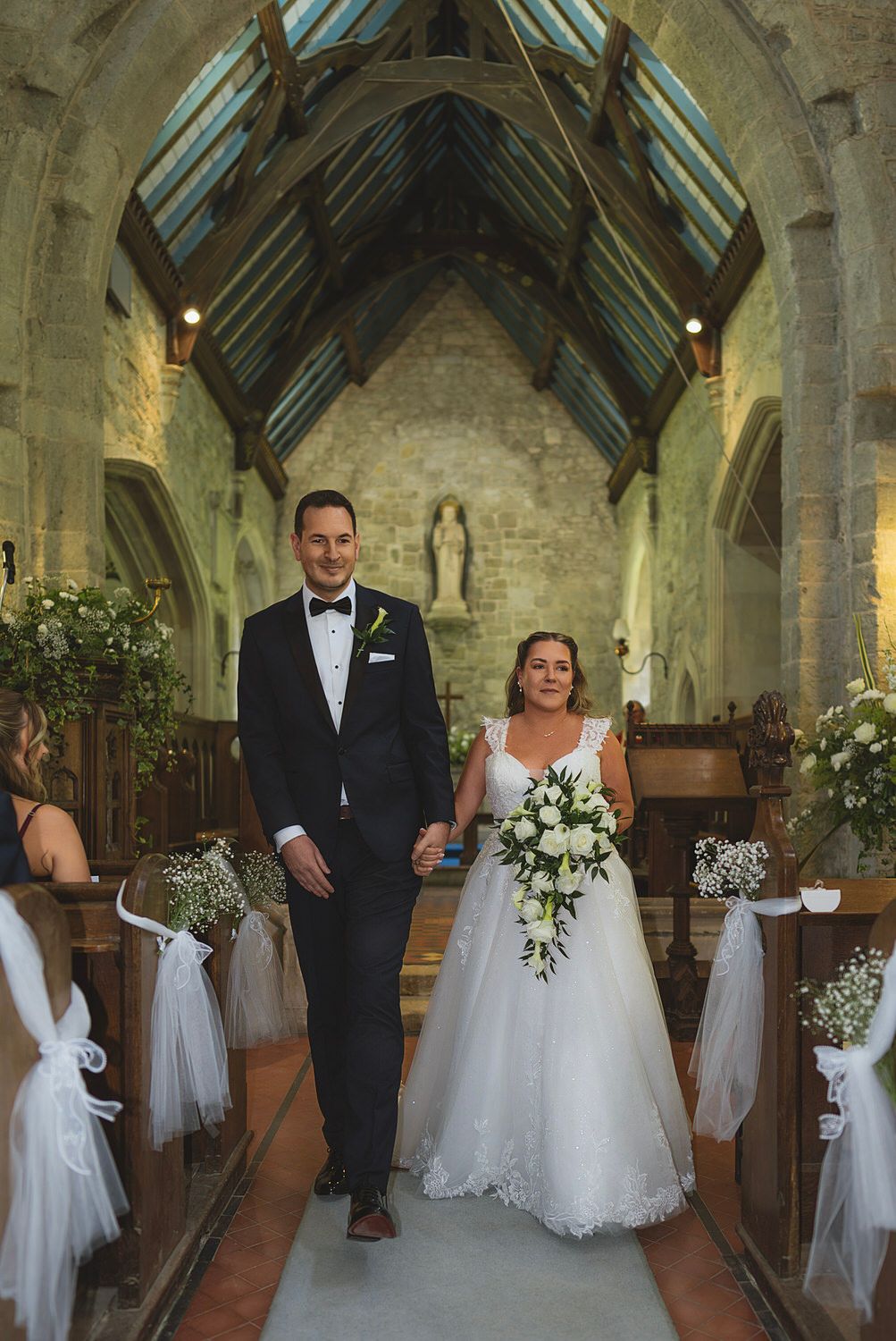 Bride and groom walk down the aisle of a stone church, bride holding white floral bouquet, pews decorated with tulle bows.