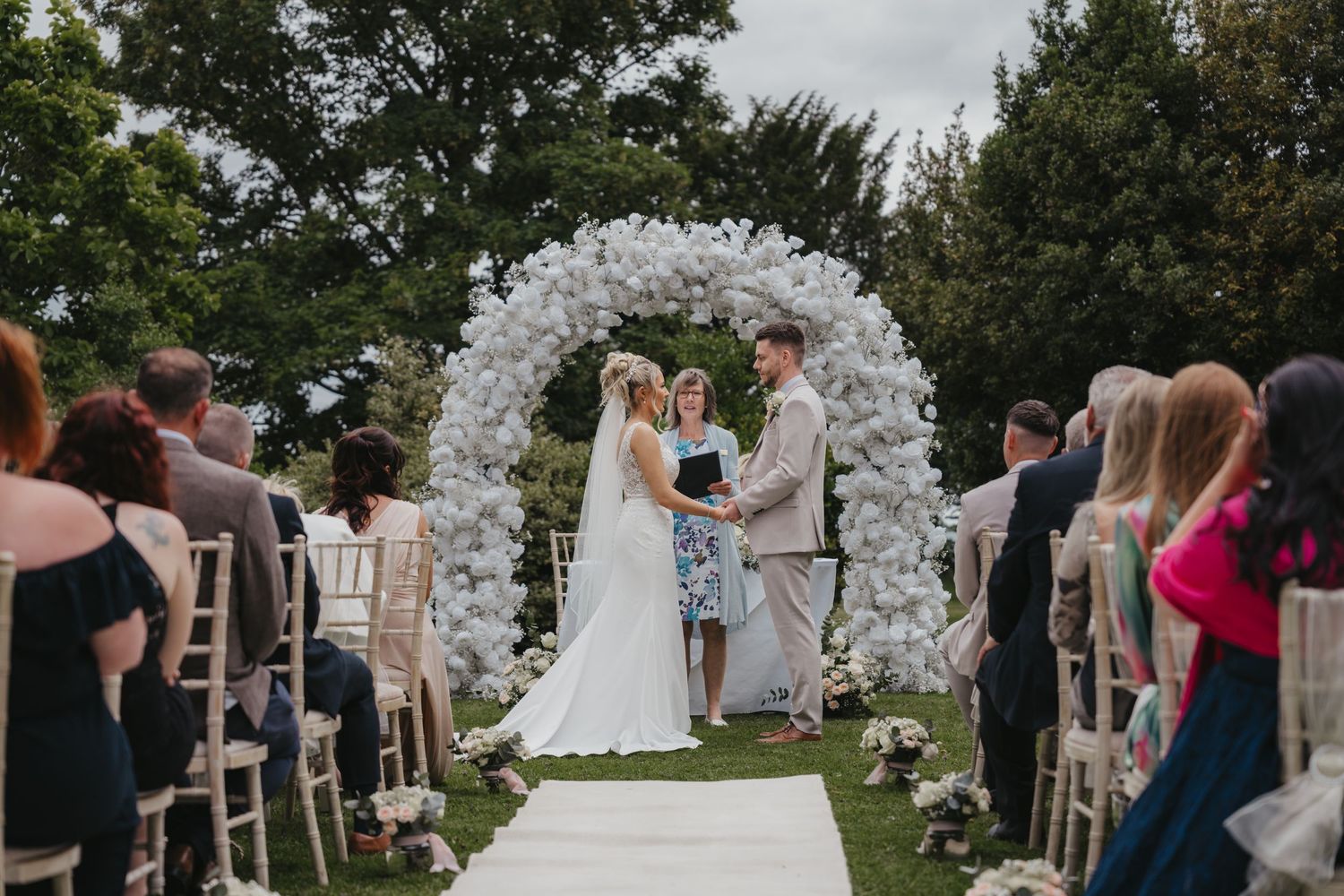 Couple exchanging vows at outdoor wedding ceremony under white floral arch, guests seated on wooden chairs.