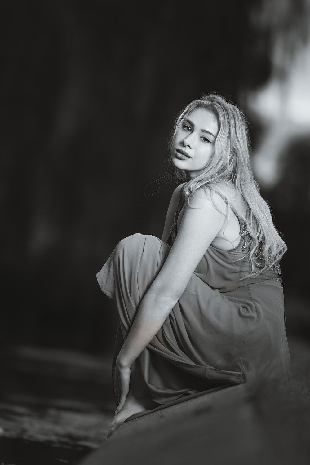 Black and white portrait of a blonde woman in a flowing dress sitting gracefully outdoors, gazing at the camera.