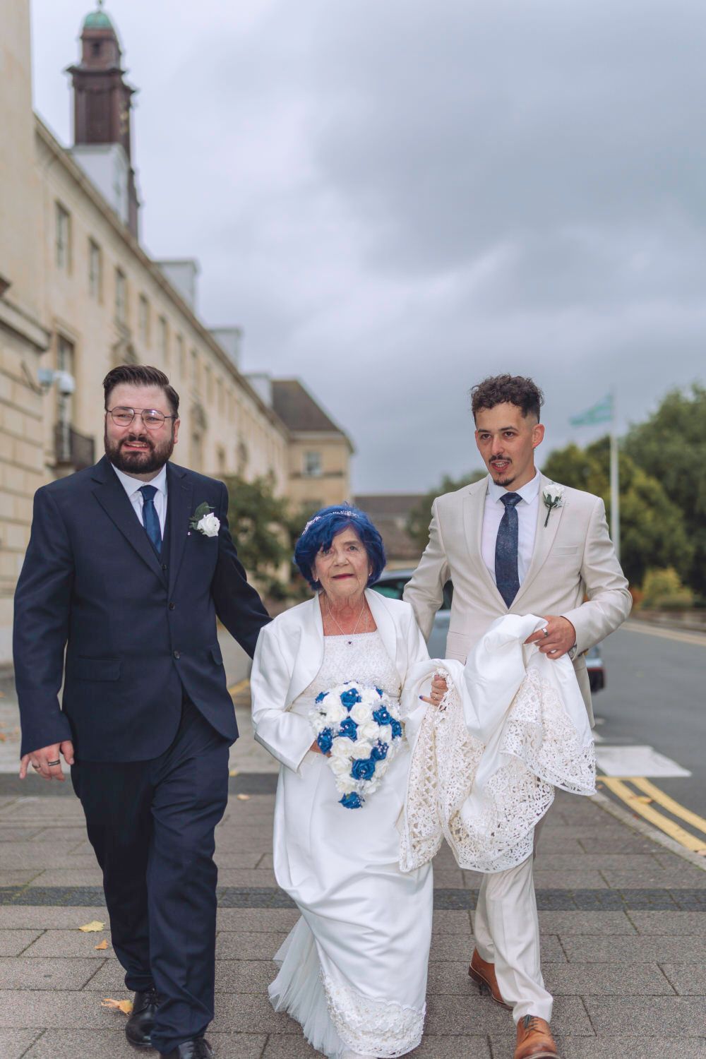Bride in white with blue bouquet walks with two suited men outside a wedding venue.