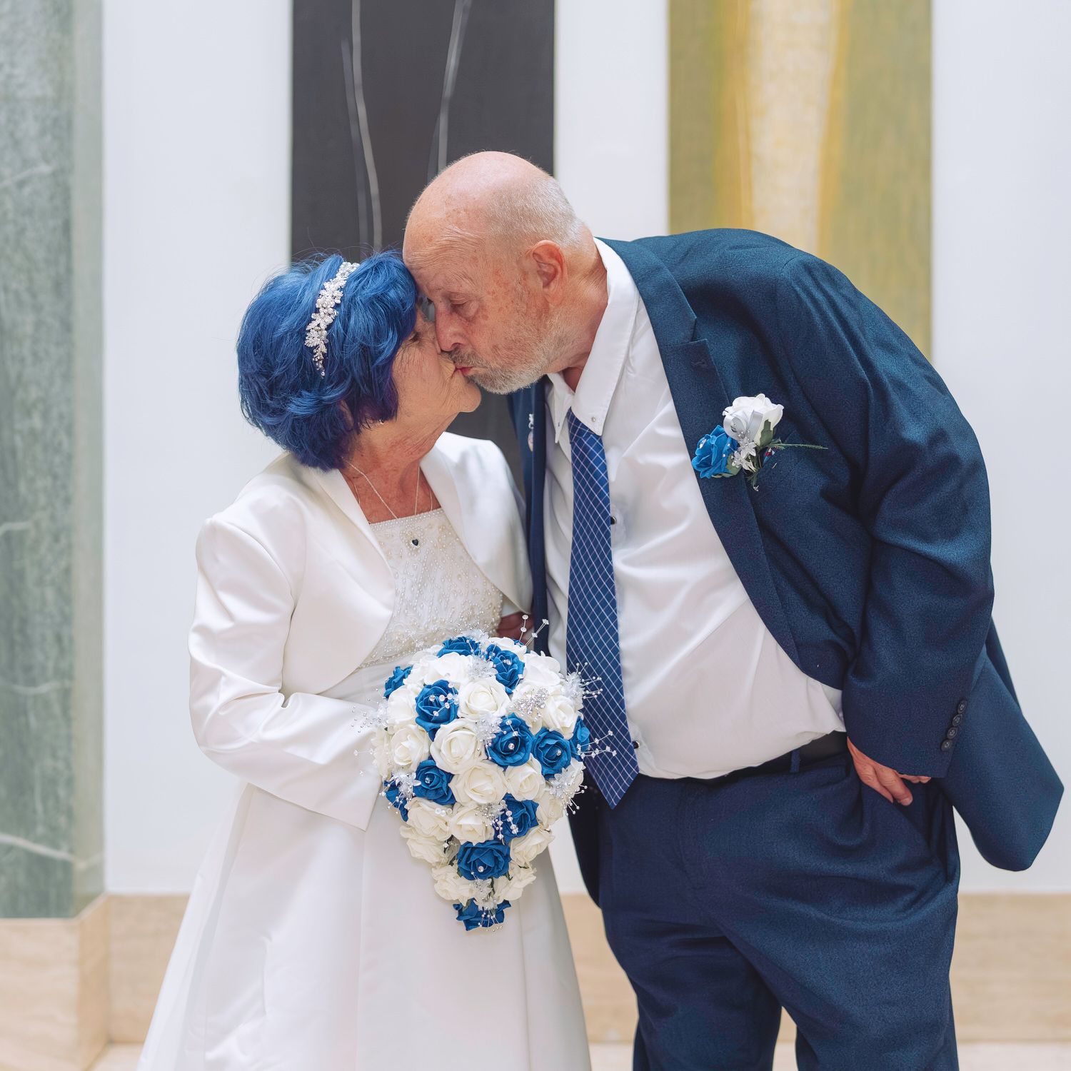 Elderly couple sharing a kiss at their wedding, bride in white with blue hair holding a blue and white bouquet.
