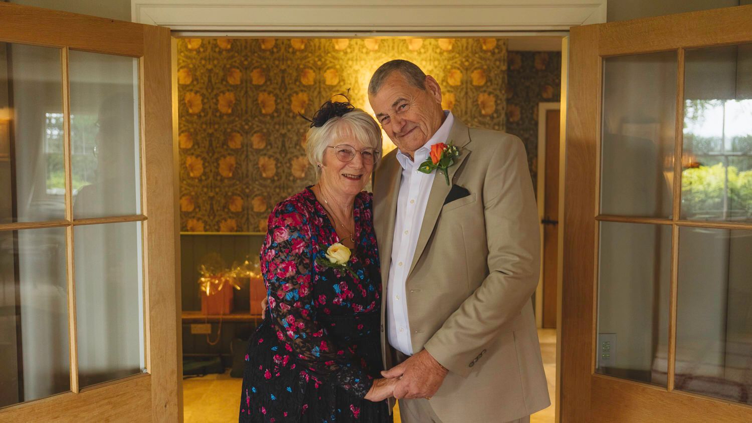 Elderly couple smiling together at a wedding, woman in floral dress and man in beige suit with boutonniere.