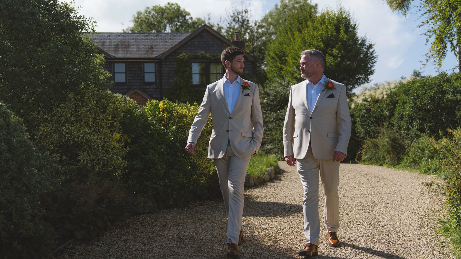 Two grooms in matching beige suits walking hand in hand along garden path.