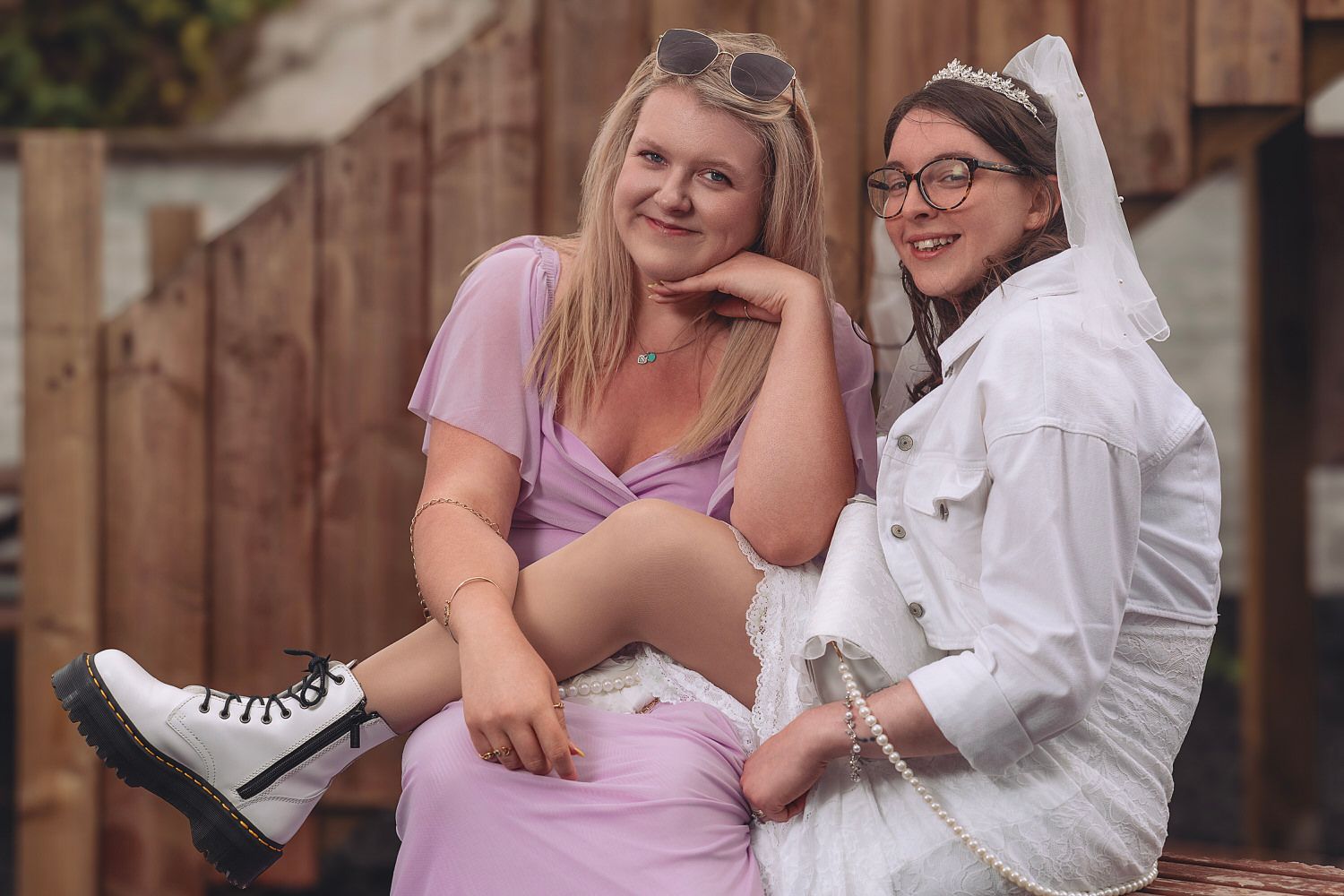 Two smiling women sitting together outdoors, one in a pink dress with white boots, the other in a white outfit with glasses.