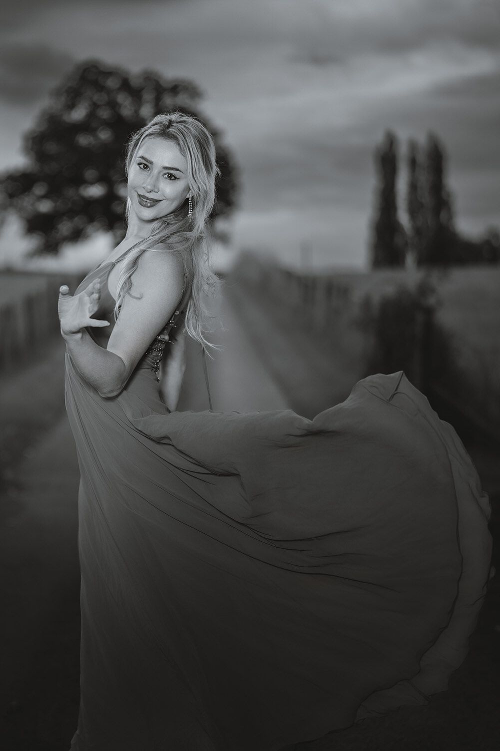 Black and white photo of a blonde woman in a flowing gown posing gracefully outdoors at dusk.