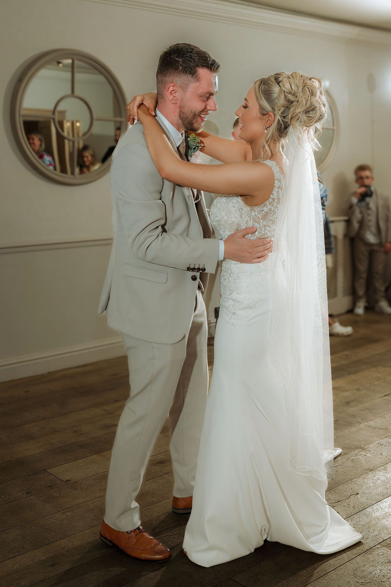 Bride and groom share their first dance, she in a white lace gown with veil, he in a light gray suit.