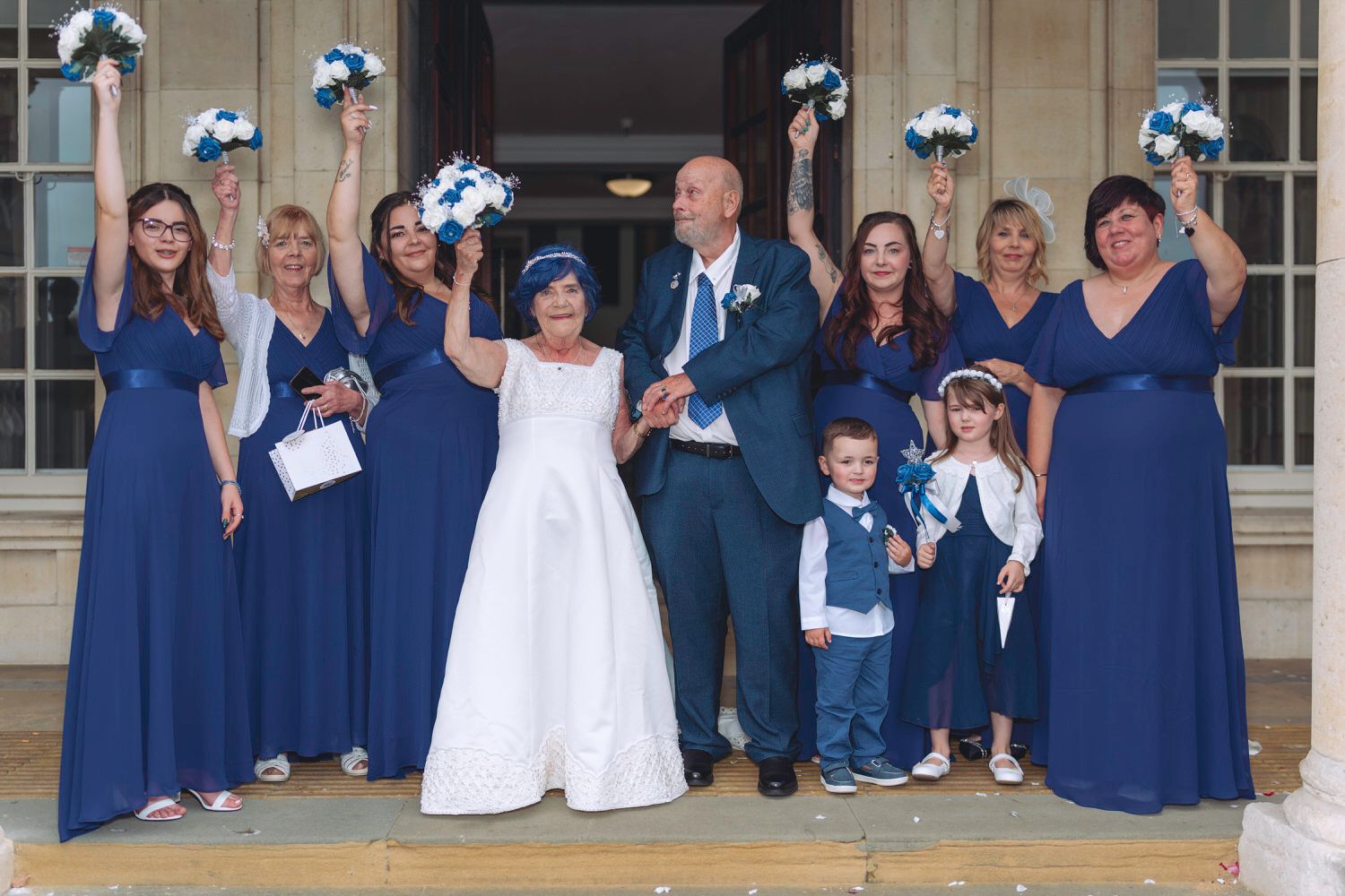 Wedding party in navy blue attire celebrating outdoors, bridesmaids raising bouquets with joyful bride and groom at center.