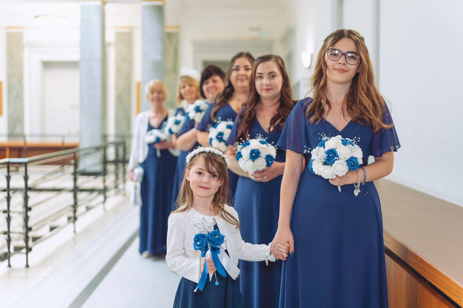 Bridesmaids in blue dresses holding teal and white bouquets stand in a line with a flower girl in a wedding hallway.