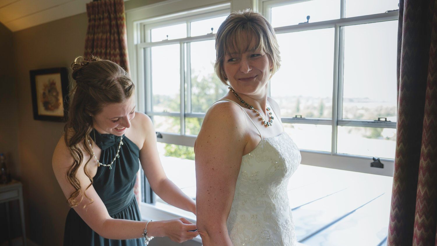 Bridesmaid in teal dress helping bride in white lace gown get dressed near a bright window on wedding day.