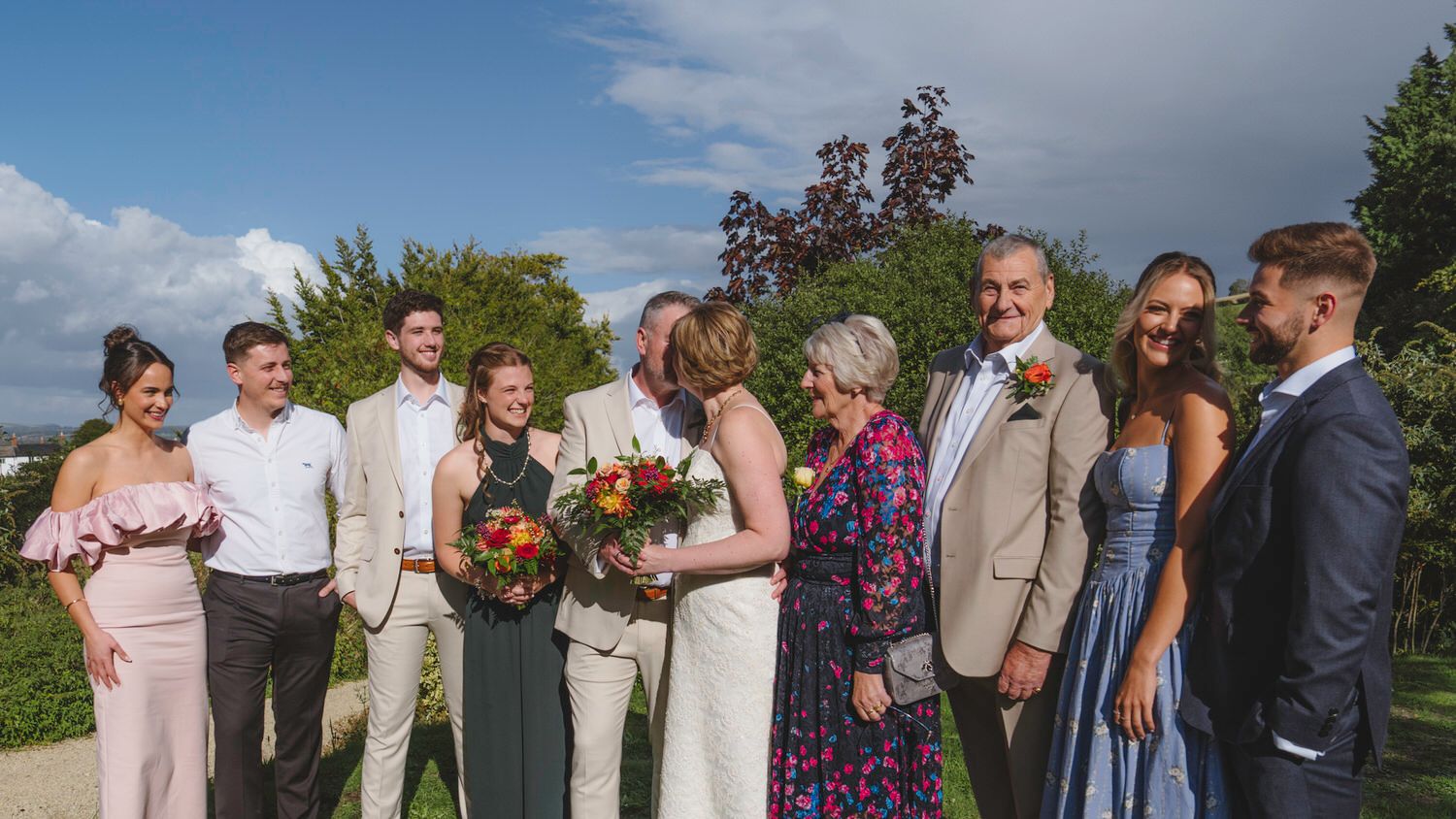Wedding party group photo outdoors with bride and groom kissing, surrounded by family in formal attire under blue sky.
