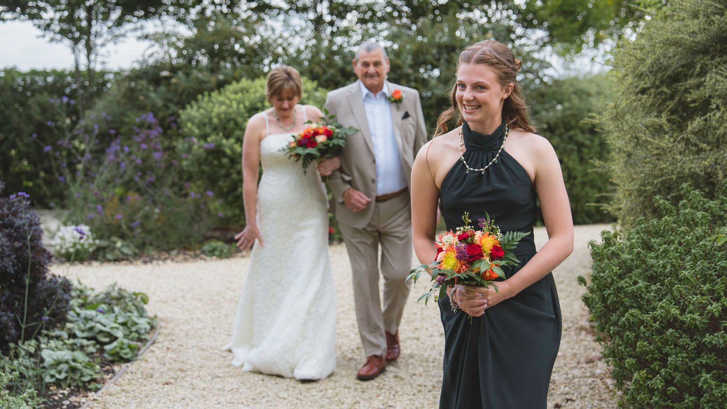 Bride in white gown walks with escort as bridesmaid in black dress leads the way through garden path.