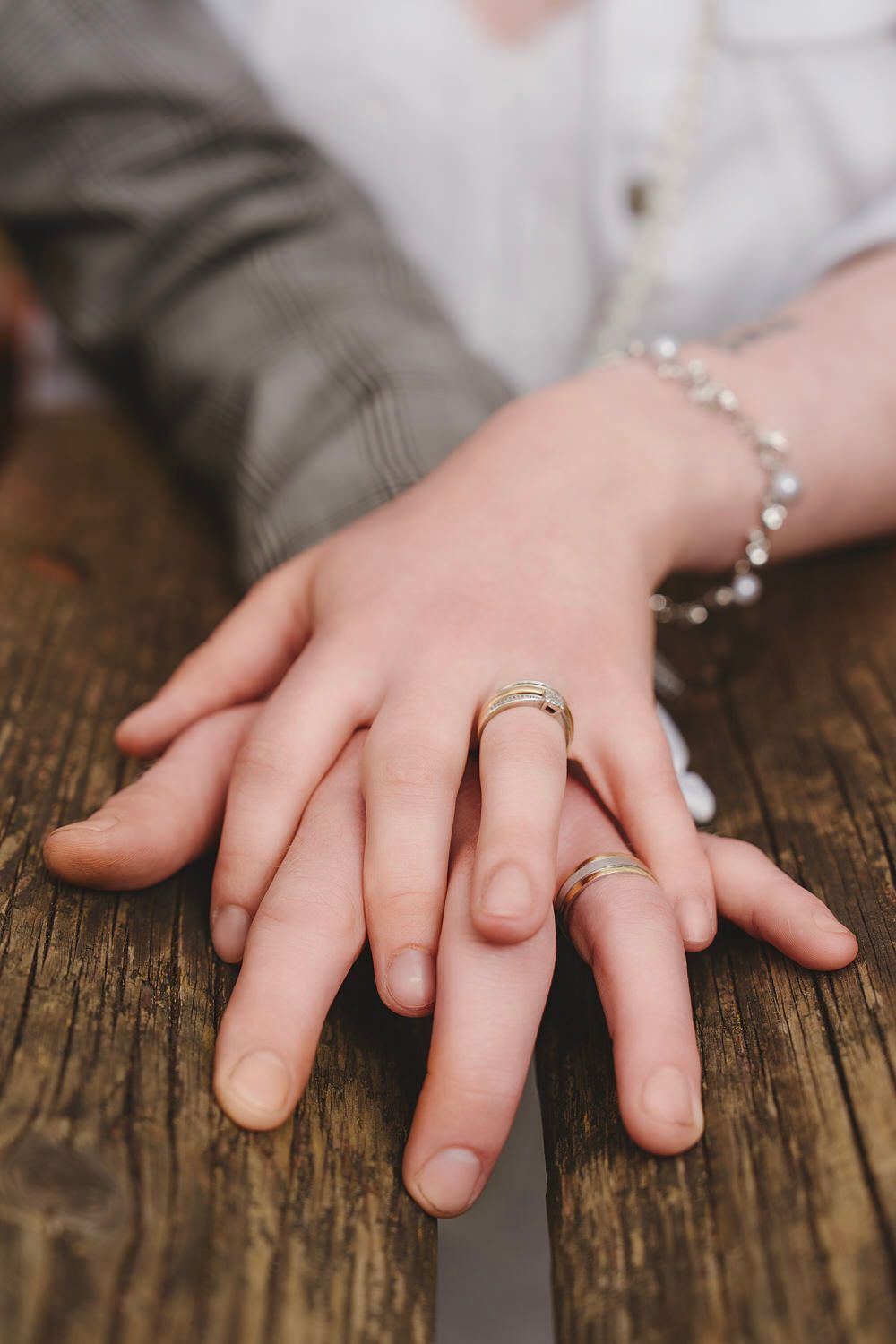 Two hands resting on wood surface displaying matching wedding bands, with a delicate bracelet visible on one wrist.