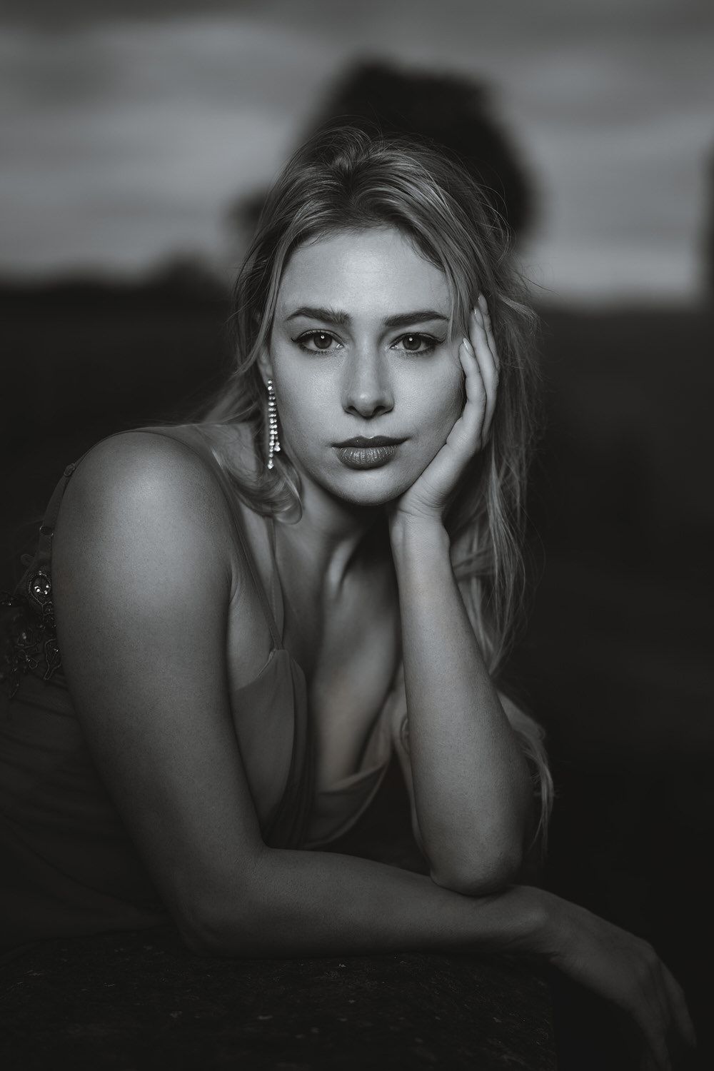 Black and white portrait of a blonde woman resting her cheek on her hand, wearing drop earrings, with a blurred outdoor background.