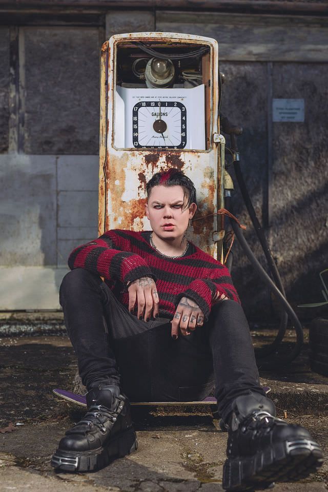 Person in red striped sweater and black platform boots sits before rusty vintage gas pump.
