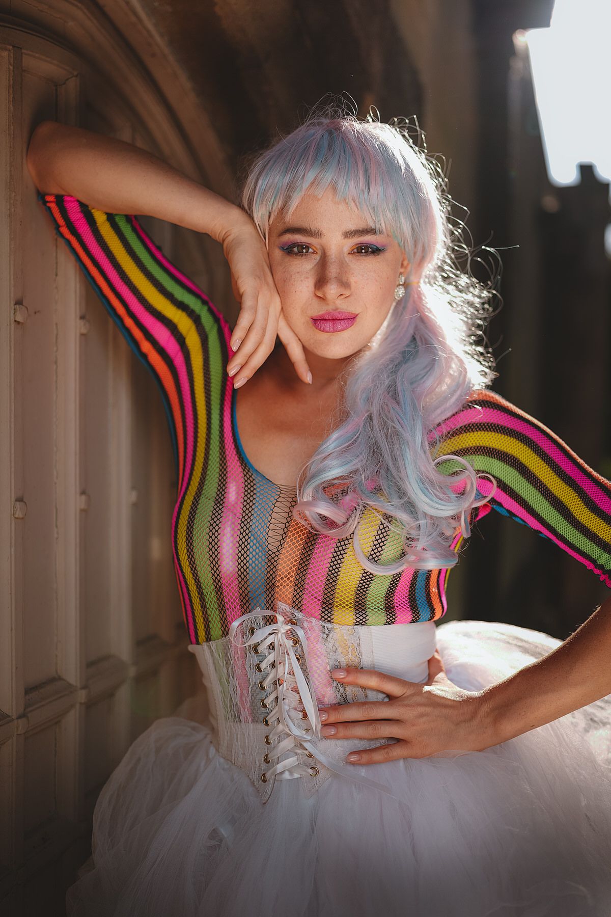 Woman with pastel lavender hair in colorful striped mesh bodysuit and white corset poses outdoors.