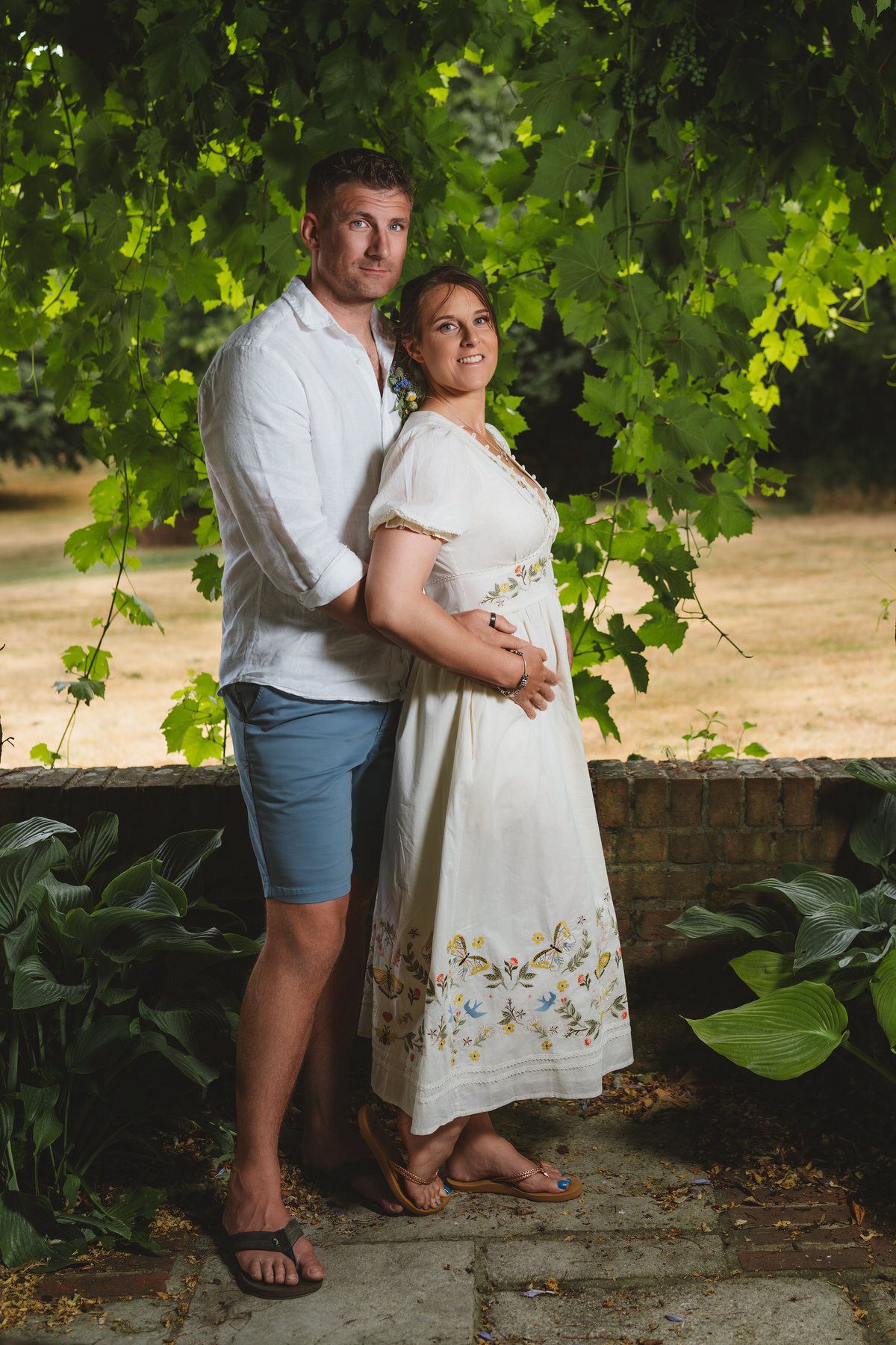 Couple embrace tenderly under lush green vine canopy in sunlit garden setting.